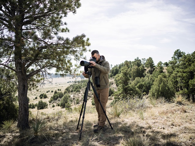 A man dressed in outdoor gear stands in a grassy, lightly wooded area, using a Swarovski spotting scope mounted on a tripod. He is focused on looking through the scope, which is set up under the partial shade of a tree. The background reveals a mix of trees, shrubs, and open landscape under a partly cloudy sky, indicating he is engaged in observing nature or wildlife.
