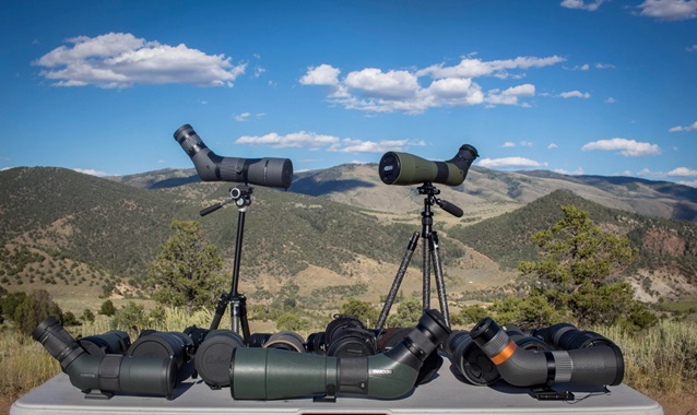 A collection of spotting scopes is displayed on a table set against a backdrop of rolling hills and a clear blue sky with scattered clouds. Two of the spotting scopes are mounted on tripods, standing upright, while several others lie on the table, showcasing different models and designs. The scenic landscape suggests a setting ideal for nature observation or birdwatching.