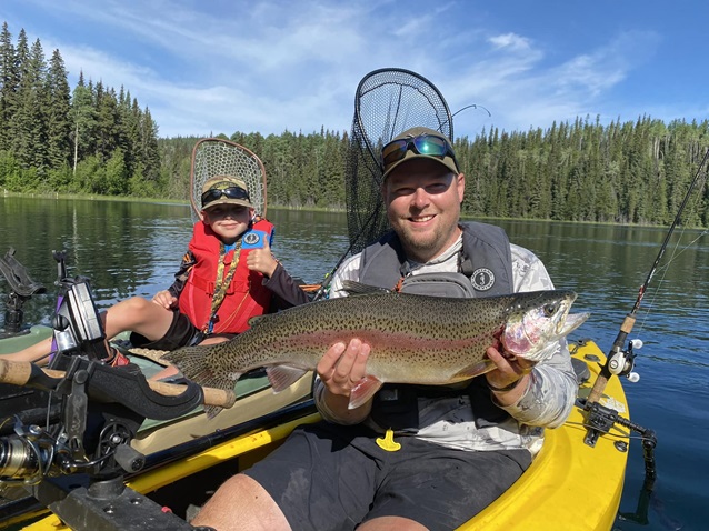 A man and a young child are sitting in kayaks on a calm lake surrounded by a forest. The man is proudly holding a large fish, while the child smiles and gives a thumbs-up. Both are wearing life jackets and have fishing equipment on their kayaks. The sky is clear, and the water reflects the surrounding trees, indicating a sunny day perfect for fishing.