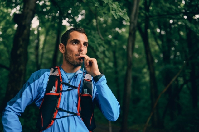 A person in a blue jacket standing in the woods, drinking from a hydration pack strapped to their back.